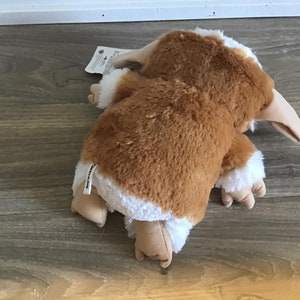 May include: A plush toy of a creature with brown fur, white accents, and a long, beige beak. The toy is lying on its stomach, with its arms and legs extended. The toy has a tag attached.