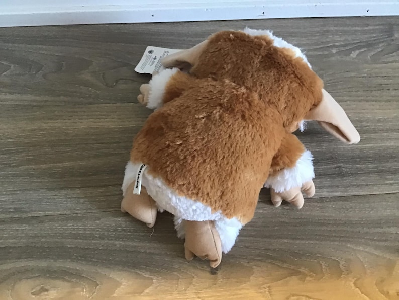 May include: A plush toy of a creature with brown fur, white accents, and a long, beige beak. The toy is lying on its stomach, with its arms and legs extended. The toy has a tag attached.