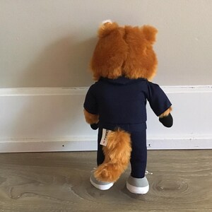 Pu&ograve; includere: Un peluche a forma di volpe, vista da dietro, indossa una tuta blu navy con cappuccio. La volpe ha la pelliccia arancione, una coda soffice e scarpe da ginnastica grigie e bianche. Il giocattolo &egrave; appoggiato a un muro bianco.