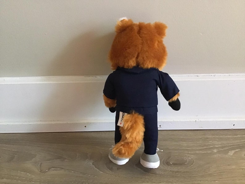 Pu&ograve; includere: Un peluche a forma di volpe, vista da dietro, indossa una tuta blu navy con cappuccio. La volpe ha la pelliccia arancione, una coda soffice e scarpe da ginnastica grigie e bianche. Il giocattolo &egrave; appoggiato a un muro bianco.