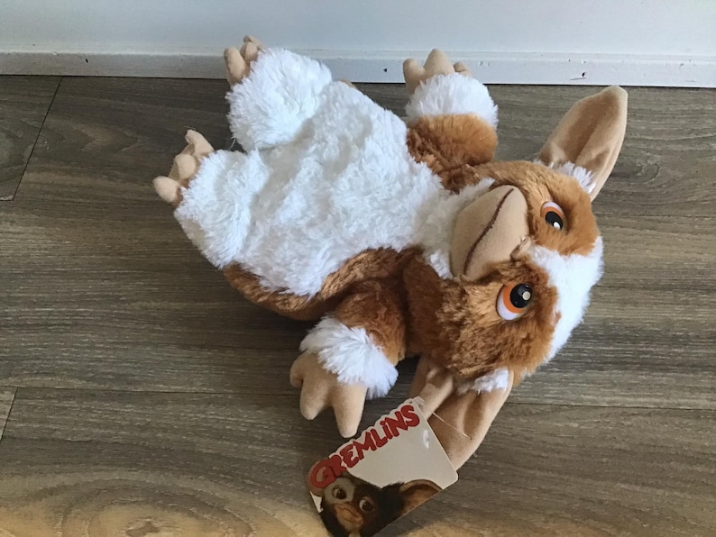 May include: A plush Gremlins character toy in brown, white, and tan. The toy has large ears, orange eyes, and a tag that reads "Gremlins". The plush toy is lying on a wooden surface.