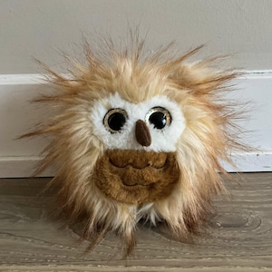 May include: A plush owl toy with a fluffy, textured appearance. The owl features large, shiny eyes, a small brown beak, and a brown chest. The toy's fur is a mix of light brown, tan, and white, giving it a soft, cuddly look.