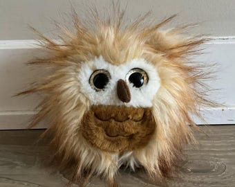 May include: A plush owl toy with a fluffy, textured appearance. The owl features large, shiny eyes, a small brown beak, and a brown chest. The toy's fur is a mix of light brown, tan, and white, giving it a soft, cuddly look.