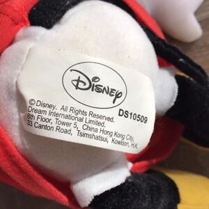 May include: A white plush toy with a red and yellow outfit. The toy has a black face and a white oval-shaped tag with the text "Disney" and "DS10509". The tag also includes copyright information and the address of Dream International Limited.
