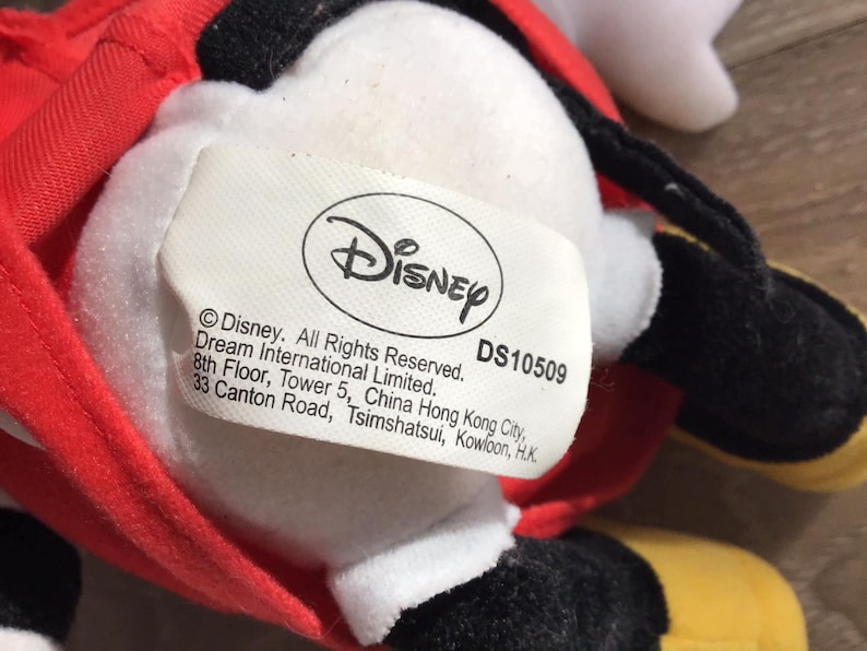 May include: A white plush toy with a red and yellow outfit. The toy has a black face and a white oval-shaped tag with the text "Disney" and "DS10509". The tag also includes copyright information and the address of Dream International Limited.