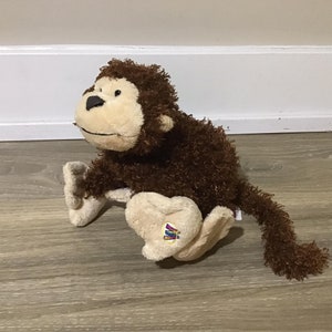 May include: A brown and tan monkey plush toy with a long tail. The monkey has brown, curly fur and a tan face with black eyes and a black nose. The monkey is sitting on its bottom with its legs outstretched in front of it. The monkey has a small, colourful tag on its foot.