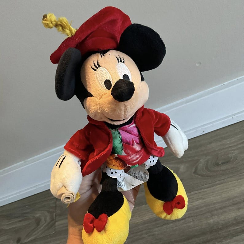 May include: A Minnie Mouse plush doll wearing a red jacket and hat. The doll has black ears, a tan face, and yellow shoes with red bows. The jacket has a floral design.