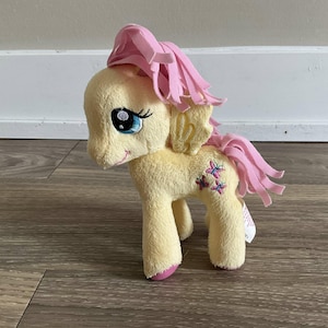 May include: A soft toy pony, predominantly yellow with pink details. The pony features a pink mane and tail, blue embroidered eyes, and a small smile. It has butterfly designs on its flank. The toy is designed to stand upright.