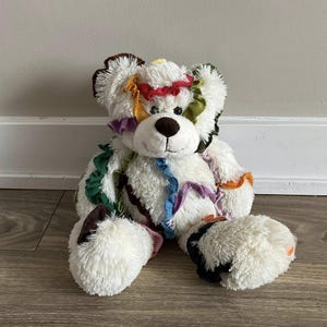 Peut inclure: Un ours en peluche blanc avec des bouts de tissu colorés cousus sur sa fourrure. L'ours a un nez brun et des yeux noirs.