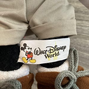May include: A plush toy wearing beige cuffed shorts and brown lace-up boots with gray laces. A white fabric tag is attached, featuring a graphic of Mickey Mouse and the black text Walt Disney World with a yellow castle silhouette in the background.