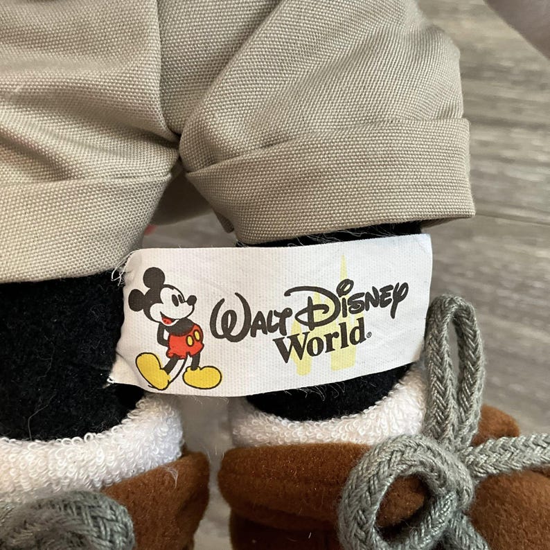 May include: A plush toy wearing beige cuffed shorts and brown lace-up boots with gray laces. A white fabric tag is attached, featuring a graphic of Mickey Mouse and the black text Walt Disney World with a yellow castle silhouette in the background.