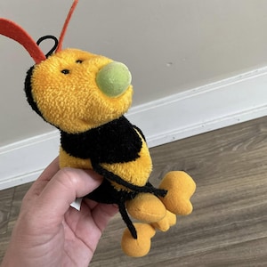 Pode incluir: Um brinquedo de pelúcia de abelha amarelo e preto com um nariz verde e antenas laranja. A abelha está sentada em uma superfície de madeira.
