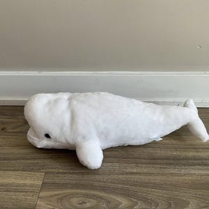 May include: A plush white beluga whale toy with a soft, textured surface. The toy has small black eyes and flipper-like appendages. The whale is lying on a wooden surface.