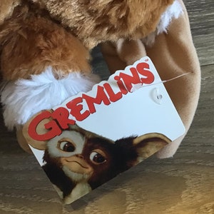 May include: A plush toy featuring a brown and white furry design, with a tag that reads "Gremlins." The tag displays a cartoon image of a Gremlin character. The toy appears soft and cuddly, with a focus on the character's face.