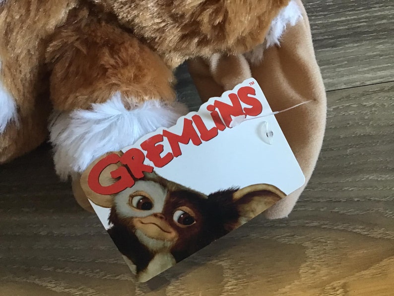 May include: A plush toy featuring a brown and white furry design, with a tag that reads "Gremlins." The tag displays a cartoon image of a Gremlin character. The toy appears soft and cuddly, with a focus on the character's face.