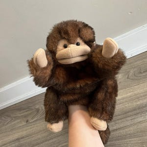 May include: A brown plush monkey puppet with a tan face, hands, and feet. The puppet has a furry texture and is designed to be worn on the hand. The monkey's arms are raised, and its eyes are visible.