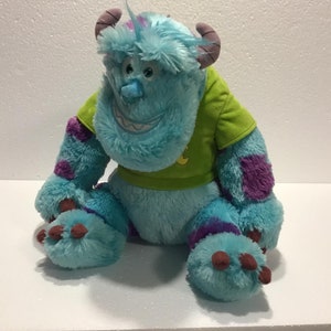 Op de afbeelding: Een blauw en paars pluche speelgoed van het personage Sulley uit de film Monsters Inc. Het speelgoed draagt een groen shirt met een geel symbool.