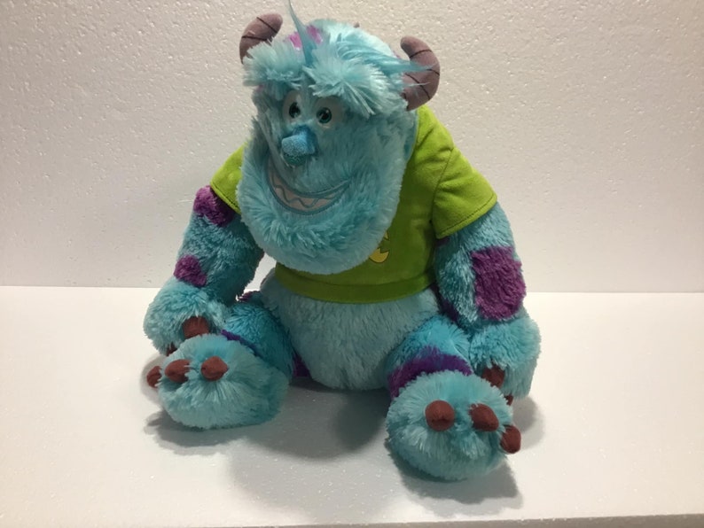Op de afbeelding: Een blauw en paars pluche speelgoed van het personage Sulley uit de film Monsters Inc. Het speelgoed draagt een groen shirt met een geel symbool.