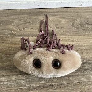 May include: A plush toy shaped like a microbe, with a light tan body and large, dark brown eyes. The toy has several purple, rope-like appendages extending from the top. The toy is designed to resemble a microorganism.