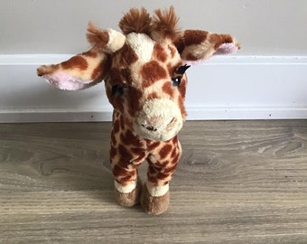 Könnte beinhalten: Ein braunes und hellbraunes Plüsch-Giraffe-Spielzeug mit schwarzen Augen und einer rosa Nase. Die Giraffe steht auf einem Holzboden.