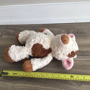 Pu&ograve; includere: Un giocattolo di peluche di mucca bianco e marrone con orecchie rosa. Il giocattolo &egrave; sdraiato su un pavimento di legno.