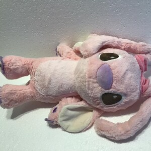 May include: Pink plush toy with purple accents and brown eyes. The toy is shaped like a cartoon character with large ears and a bow.