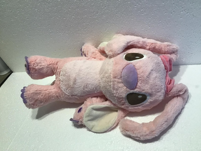 May include: Pink plush toy with purple accents and brown eyes. The toy is shaped like a cartoon character with large ears and a bow.