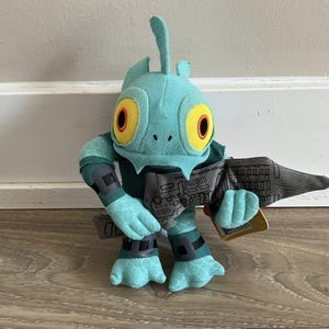 May include: A teal plush toy creature with large yellow eyes and a fin on its head. It has grey accents on its arms, legs, and a weapon-like accessory. The toy is standing on a light brown surface.