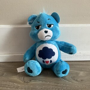 Puede incluir: Un oso de peluche azul con una expresión triste. El oso tiene una barriga blanca con un diseño de nube azul oscuro y detalles de corazón rojo. Detalles en forma de corazón en los pies del oso. Una pequeña etiqueta está adjunta.