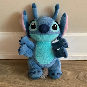 May include: A blue plush toy character with four arms, large ears with pink inner lining, two antennae, and a light blue chest patch. The character has large black eyes, a dark blue nose, and embroidered claw details on its hands and feet.