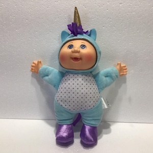 May include: A blue plush Cabbage Patch Kid doll wearing a unicorn costume with a gold horn, purple ears, and white polka dots. The doll has a pink face, blue eyes, and a big smile.