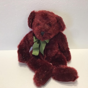 May include: A plush burgundy teddy bear with a green bow. The bear is sitting with its legs crossed and has a soft, fuzzy texture.