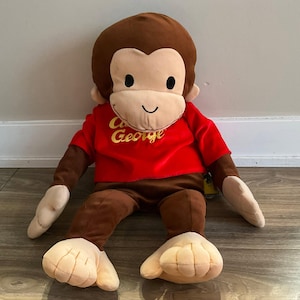 May include: A plush Curious George monkey toy featuring a brown body, light beige face, hands, and feet, and black embroidered eyes and mouth. The monkey wears a bright red short-sleeved shirt with the text Curious George embroidered in yellow.