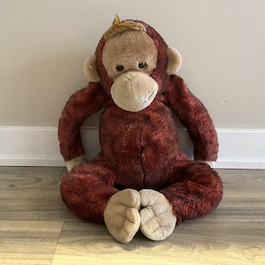 以下が含まれることがあります： 赤茶色の体、ベージュの顔、耳、足を持つ、ぬいぐるみオランウータンのおもちゃ。 ぬいぐるみは木の床に座っており、腕と脚を広げています。 おもちゃはフレンドリーな表情をしています。