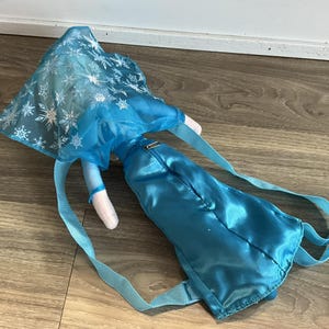May include: A blue backpack shaped like a doll, featuring a blue dress and a cape with silver snowflake patterns. The backpack has blue straps and a zipper closure. The doll's arms are white.