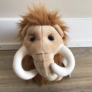 以下が含まれることがあります： 大きな牙を持つ、茶色と白のふわふわのマンモスのおもちゃ。おもちゃは茶色の毛皮で覆われた体と、黒い目をした白い顔をしています。