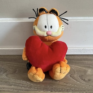 Op de afbeelding: Een pluche Garfield-speelgoed, oranje met zwarte strepen en een wit gezicht, met een groot rood hart. Het knuffeldier heeft een roze neus en zwarte snorharen. Het speelgoed staat op een houten oppervlak.