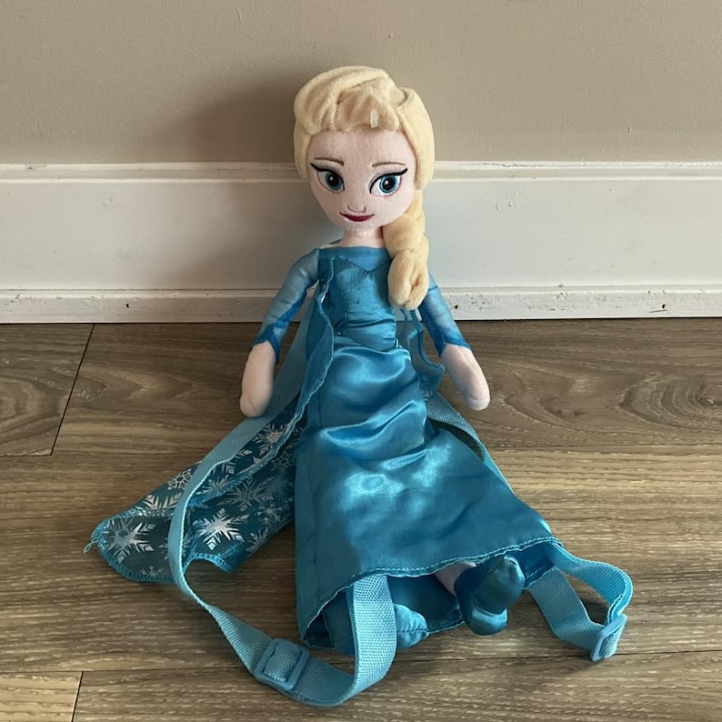 May include: A plush doll of a character with blonde hair in a side braid, wearing a blue satin dress with a cape and backpack straps. The cape has a snowflake pattern. The doll is on a wooden surface.