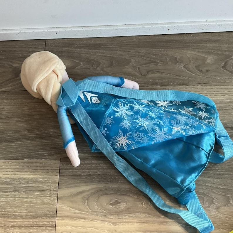 May include: A plush doll resembling a character from the movie Frozen, with a light blonde bun and blue dress. The doll has a blue backpack with snowflake patterns and blue straps.