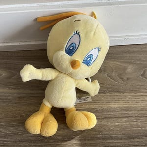 Puede incluir: Juguete de peluche amarillo Tweety Bird con ojos azules y un pico amarillo. El juguete tiene una pequeña etiqueta en su pierna con texto en varios idiomas.
