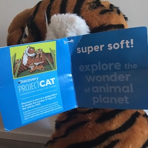 May include: A plush tiger toy with orange and black stripes. The toy is holding a blue tag that says "Discovery Project Cat Conserving Acres for Tigers". The tag also has a picture of a tiger family. The tag on the other side says "super soft! explore the wonder of animal planet".