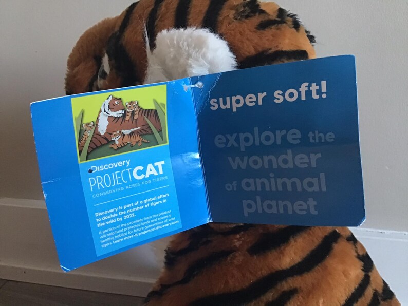 May include: A plush tiger toy with orange and black stripes. The toy is holding a blue tag that says "Discovery Project Cat Conserving Acres for Tigers". The tag also has a picture of a tiger family. The tag on the other side says "super soft! explore the wonder of animal planet".