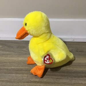 May include: A yellow plush duck with an orange beak and feet. The duck has a Ty tag attached to its side.