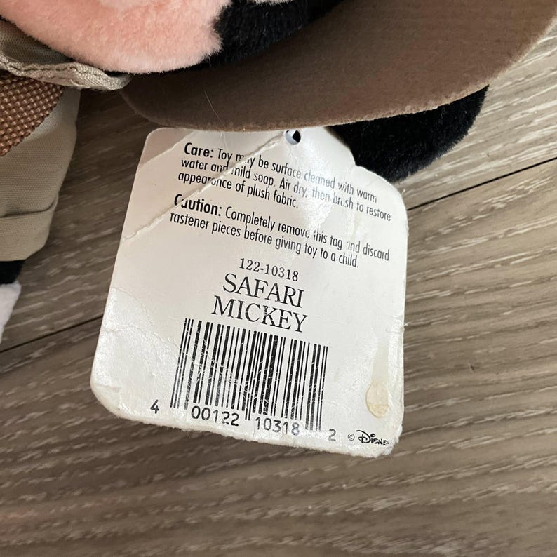 May include: A plush Safari Mickey toy with a white paper tag attached. The tag features the text 'SAFARI MICKEY', a barcode, and care instructions. The toy has soft black, pink, and tan fabric components visible, including a brown brimmed hat.