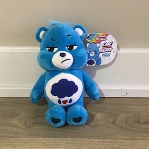May include: Blue Care Bear plush toy with a grumpy expression and a dark blue cloud with raindrops and a red heart on its belly. The Care Bears logo is on a tag attached to the toy.
