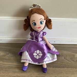 Puede incluir: Muñeca de peluche de una princesa de dibujos animados, con cabello castaño, ojos azules y un vestido morado con detalles blancos. La muñeca tiene una tiara plateada y zapatos morados. El vestido tiene un diseño de flor blanca.