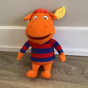 May include: A plush toy of a cartoon moose with orange skin, a red and blue striped shirt, and yellow antlers. The character has a wide, dark brown smile, white eyes with black pupils, and a soft, stuffed texture throughout its entire body.