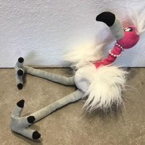 Può includere: Un giocattolo di peluche di fenicottero rosa e bianco con una collana di perle. Il fenicottero ha un collo lungo e gambe lunghe con piedi neri. Il giocattolo è sdraiato sul fianco.