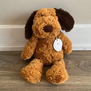 May include: A plush dog toy with soft, textured golden-brown fur and dark brown ears. The stuffed animal features a dark brown nose and a round white tag attached to its neck that reads The Manhattan Toy Company with a small colorful logo.