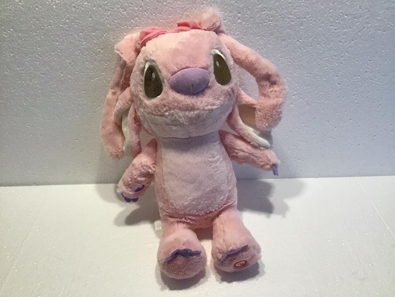 May include: Pink plush toy with large ears and a pink bow. The toy has brown eyes, a purple nose, and purple feet.